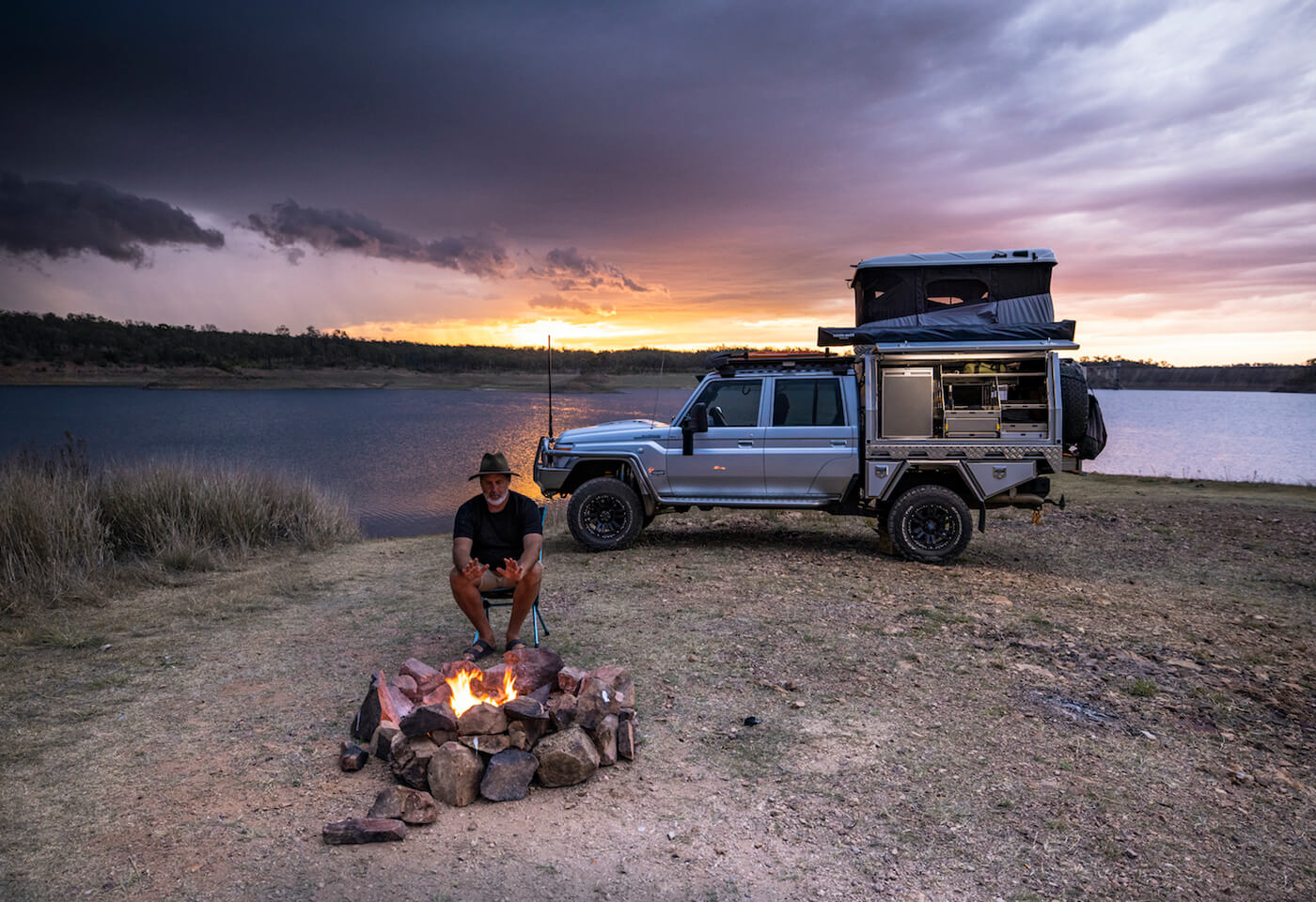 Image of man with a hat warming hands by fire in front of a 4WD.